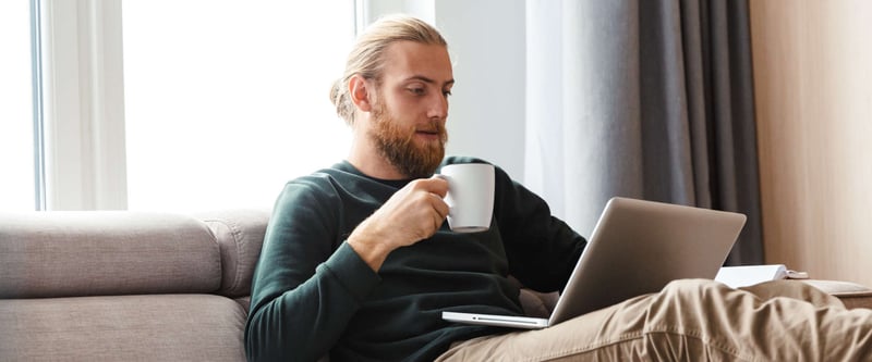 male drinking coffee on a couch while using a laptop computer